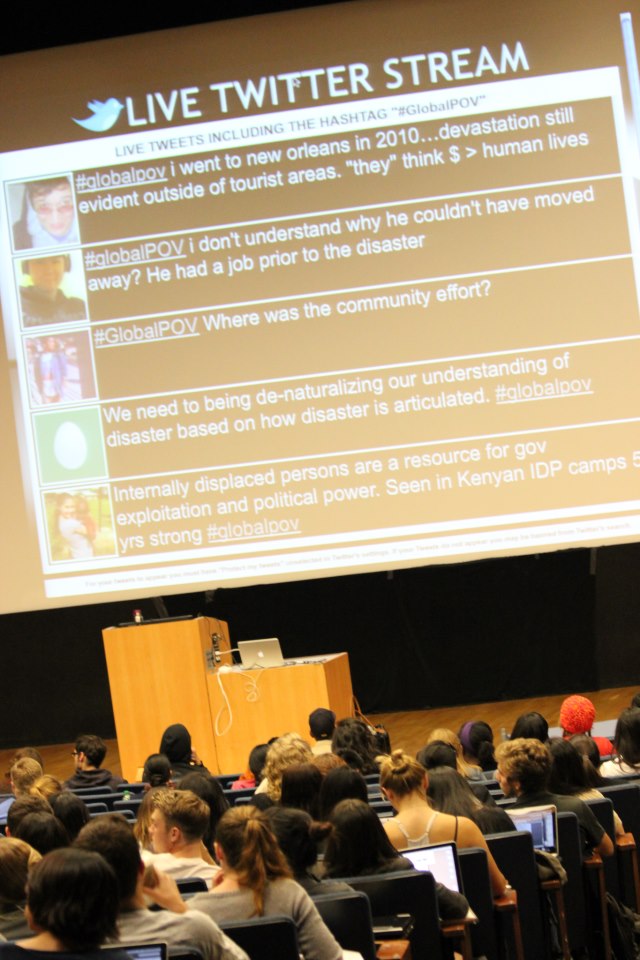 A large lecture hall filled with students, most of whom are using laptops. At the front of the room, there is a large screen displaying a live Twitter stream with tweets related to the hashtag "#GlobalPOV." The tweets cover various topics, including reflections on disaster recovery, community efforts, and the importance of addressing the needs of internally displaced persons.