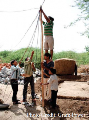 A group of five men is working together to install an electricity pole in a rural area. One man stands on a large rock, securing the pole at the top, while the others support and stabilize it from below. 