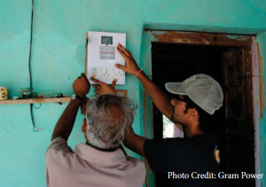 Two men are installing an electrical device, possibly a power meter, on a turquoise-painted wall indoors.