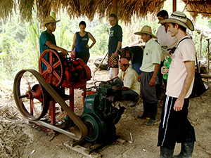 A group of people gathered under a thatched roof structure, observing a mechanical setup. The setup includes a large red and green engine with belts and pulleys. The group consists of men and women, some wearing hats and outdoor clothing.