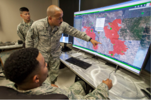 Three military personnel in uniform are in a command center. One individual is pointing at a large screen displaying a detailed map with highlighted areas in red, likely indicating zones of interest or concern. Another person is seated at a computer, using a mouse to navigate the map. The third person stands nearby, observing.