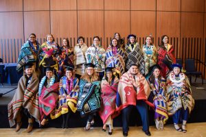 A group of graduates pose for a group photo in a large hall. They are wearing graduation caps and are wrapped in colorful, patterned blankets.