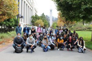 A large group of individuals, all wearing face masks, are gathered for a group photo outdoors on a university campus with the campanile in the background.