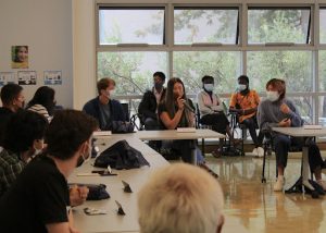 A group of people are seated around a large table in a room with floor-to-ceiling windows. Most of them are wearing face masks. Abigail Chin in the center is holding a microphone, appearing to speak to the group. Other attendees are listening attentively.