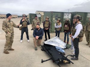 A group of people, both men and women, are gathered outdoors, likely on a military base. They are dressed in military fatigues and tactical gear, including helmets and vests. One man in uniform is standing and appears to be instructing or briefing the group.