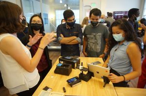 A group of young adults, both men and women, are gathered around a table in an indoor setting. They are all wearing face masks. On the table, there are various technical devices and equipment, including a tablet and what looks like a microscope or similar apparatus. A woman on the left is speaking and gesturing with her hands, possibly explaining something to the group. The group appears engaged and attentive to the explanation.