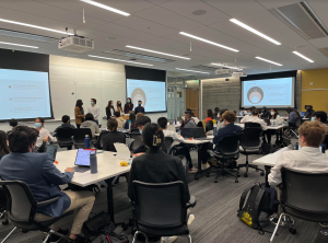 A modern classroom with rows of students seated at tables, listening to a presentation. Six presenters stand at the front of the room, each wearing masks. Two large screens display slides with market size data, including terms like "Total Addressable Market (TAM)" and "Serviceable Addressable Market (SAM)." The students are engaged, taking notes on laptops and tablets.