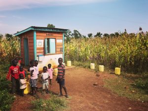 A group of children are gathered near a small, colorful building labeled "Mboto Kaka-Koka Water Project Implemented by KWAHO" in a rural area. The building has a blue door and signboard with various instructions. Several yellow water containers are scattered around the area.