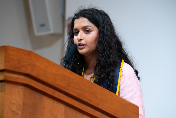 Alisha Dalvi speaking at a podium, wearing a graduation stole with the University of California, Berkeley colors.