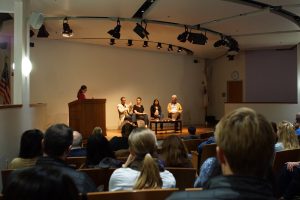 A photograph of a panel discussion taking place in an auditorium. The panel consists of four individuals seated on a stage with a moderator standing at a podium to the left. The audience, consisting of multiple people, is seated in rows facing the stage. The room is lit with spotlights on the panelists, and the background features plain walls and a small American flag on the left.