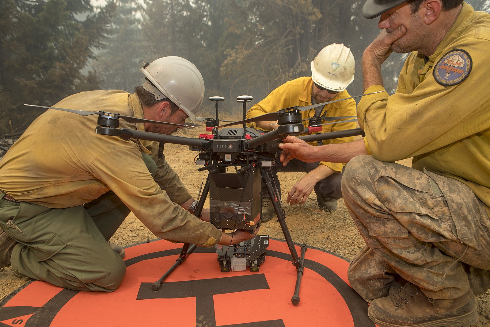 Three firefighters are gathered around a large drone on a red landing pad in a forested area. They are wearing yellow fire-resistant shirts, hard hats, and protective gear. One firefighter is making adjustments to the drone while another is observing, and a third firefighter is kneeling and contemplating the setup.