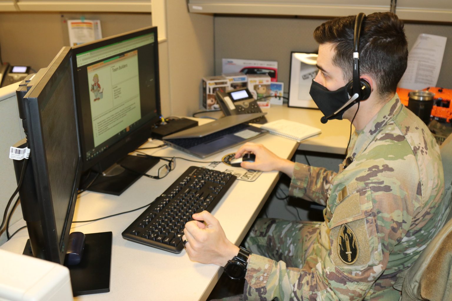 A soldier in military camouflage is sitting at a desk in an office cubicle, wearing a headset and a black face mask. He is working on a computer with dual monitors. One of the monitors shows a webpage titled "Team Builders." The desk also has a phone, a notepad, a laptop, and some decorative items.