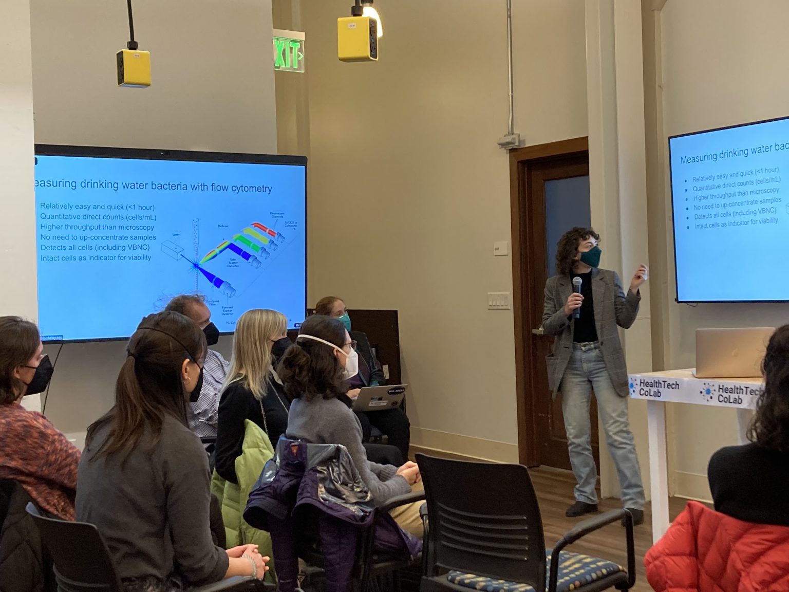 A presenter, wearing a mask, stands beside a screen displaying a slide titled "Measuring drinking water bacteria with flow cytometry." The slide includes a diagram and bullet points explaining the process. The audience, also masked, is seated and attentively listening to the presentation.