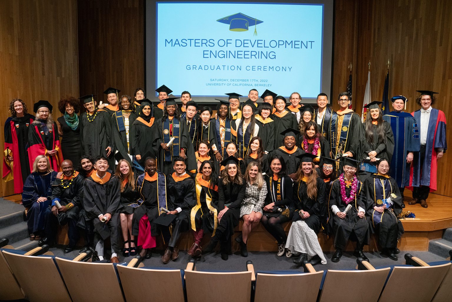 The M.DevEng Class of 2022 graduation picture. All students and faculty in graduation gown smiling for a picture. The projector screen on the background displays "Master of Development Engineering Graduation Ceremony"