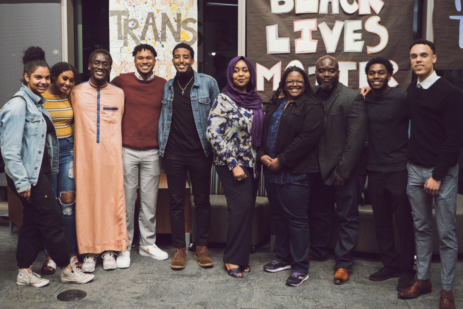 A group of ten people is standing together, smiling for a photo. They are in a room with posters on the wall that read "TRANS" and "BLACK LIVES MATTER."