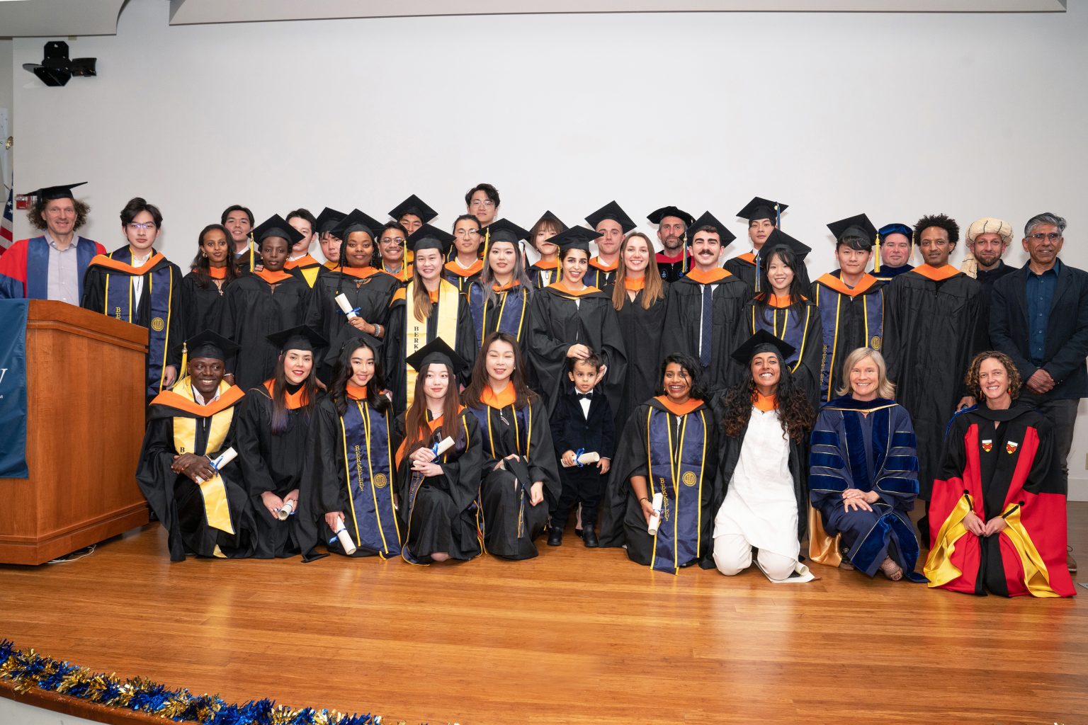 A large group of graduates poses for a photo on stage. They are all dressed in black graduation gowns and caps.