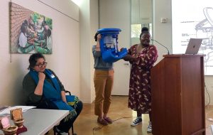 Joyce Kisiagani, with the help of another person, holds up their prototype of blue water filtration device (an affordable chlorination device that allows under-resourced communities to disinfect water at the point of collection) while giving a presenattion.