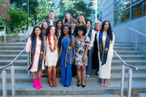 A group of 14 diverse students stands on a staircase, posing for a graduation photo. The students are smiling and wearing graduation stoles with "BERKELEY" and "CLASS OF 2024" written on them. The group includes individuals in various styles of formal attire, with some wearing leis. The background shows an outdoor setting with trees and a building with large windows.